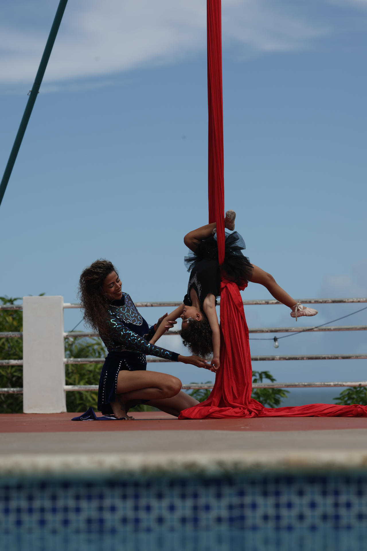 Jennifer Rivera spotting her daughter on red aerial silks outdoors, guiding her through an inverted pose with trust and joy