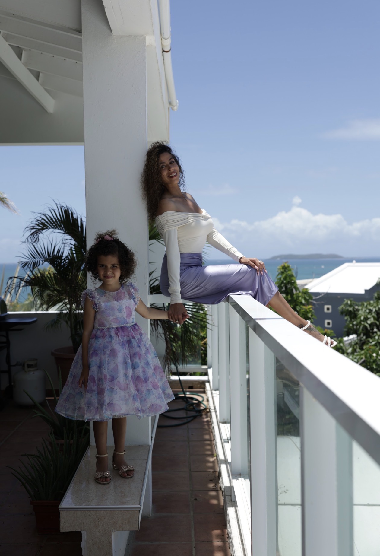 Jennifer Rivera gently holding her daughter's hand as they look out over the ocean from a bright coastal balcony.
